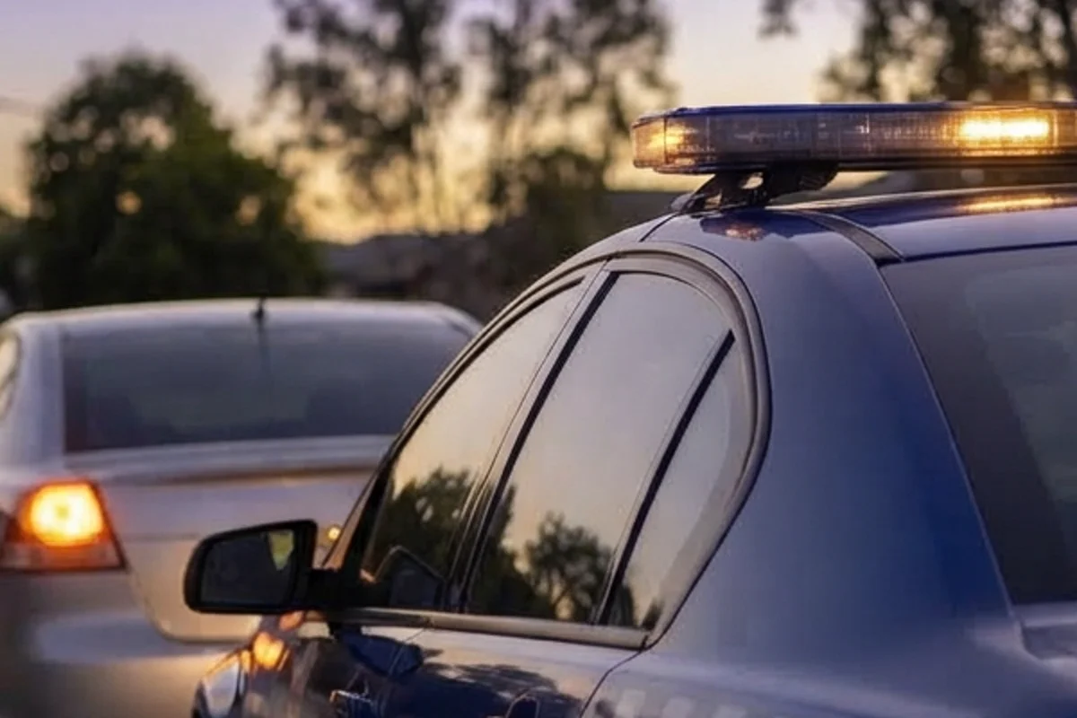 A sedan indicating left and pulling into a safe roadside verge at dusk with police lights reflected softly in the wing mirror