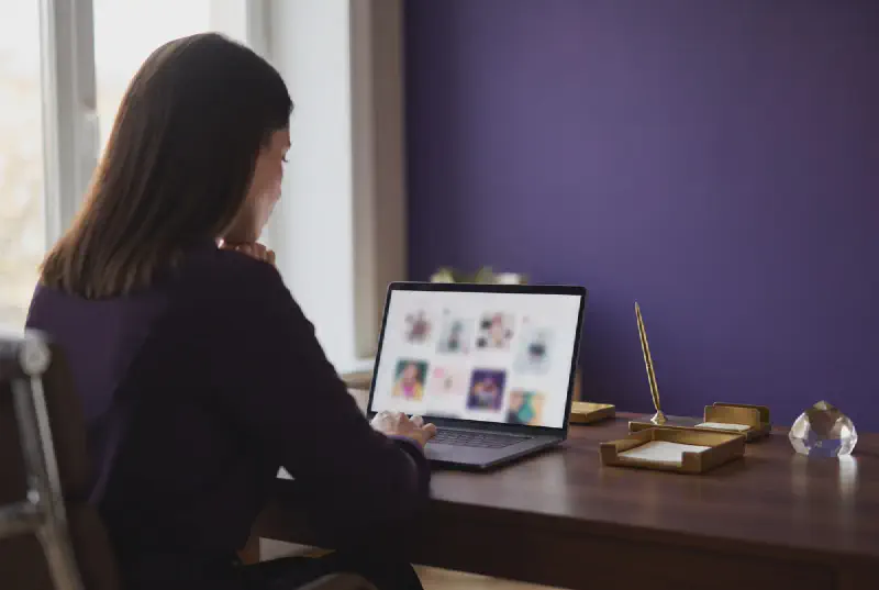 Office manager seen from behind reviewing a laptop showing blurred abstract social media tiles with royal purple wall and gold desk accessories