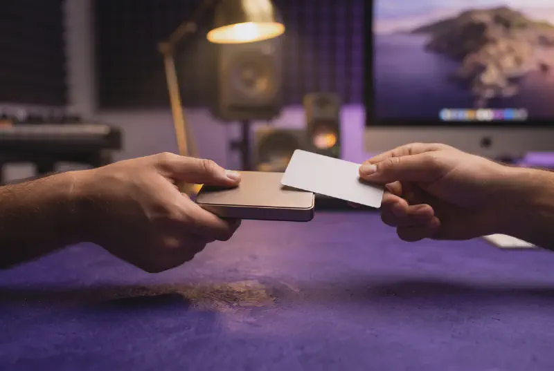 Two hands passing a small external hard drive and a blank key card across a royal purple table with gold lamp light and blurred studio background
