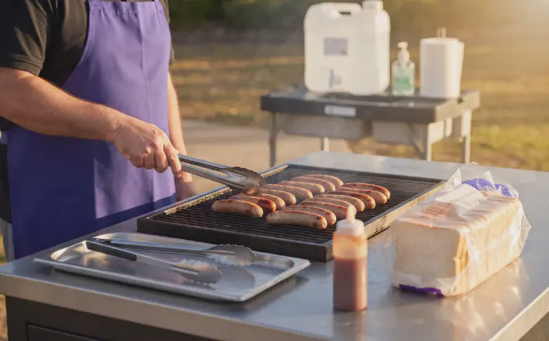 Sausages cooking at a tidy community barbecue with handwashing facilities and purple and gold details.