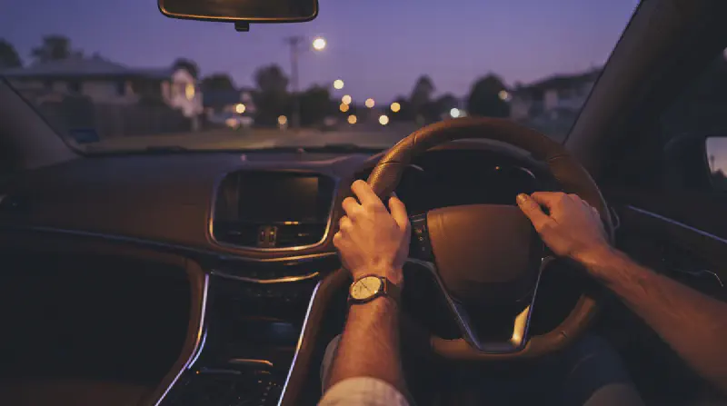 A driver's hands resting calmly on a steering wheel with the window lowered at dusk, soft purple and gold light across the dashboard