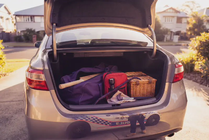 A cricket bat and a sports bag resting in an open car boot in a suburban driveway, lit in warm gold with purple accents